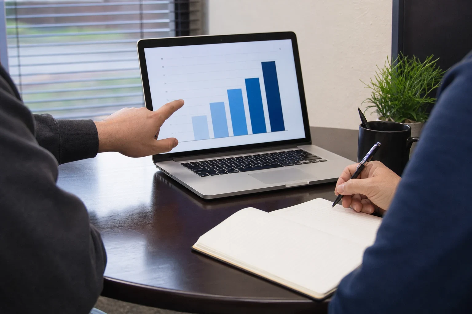 One person points at a bar chart on a laptop while another holds a pen over an open notebook at a dark round office table by a window