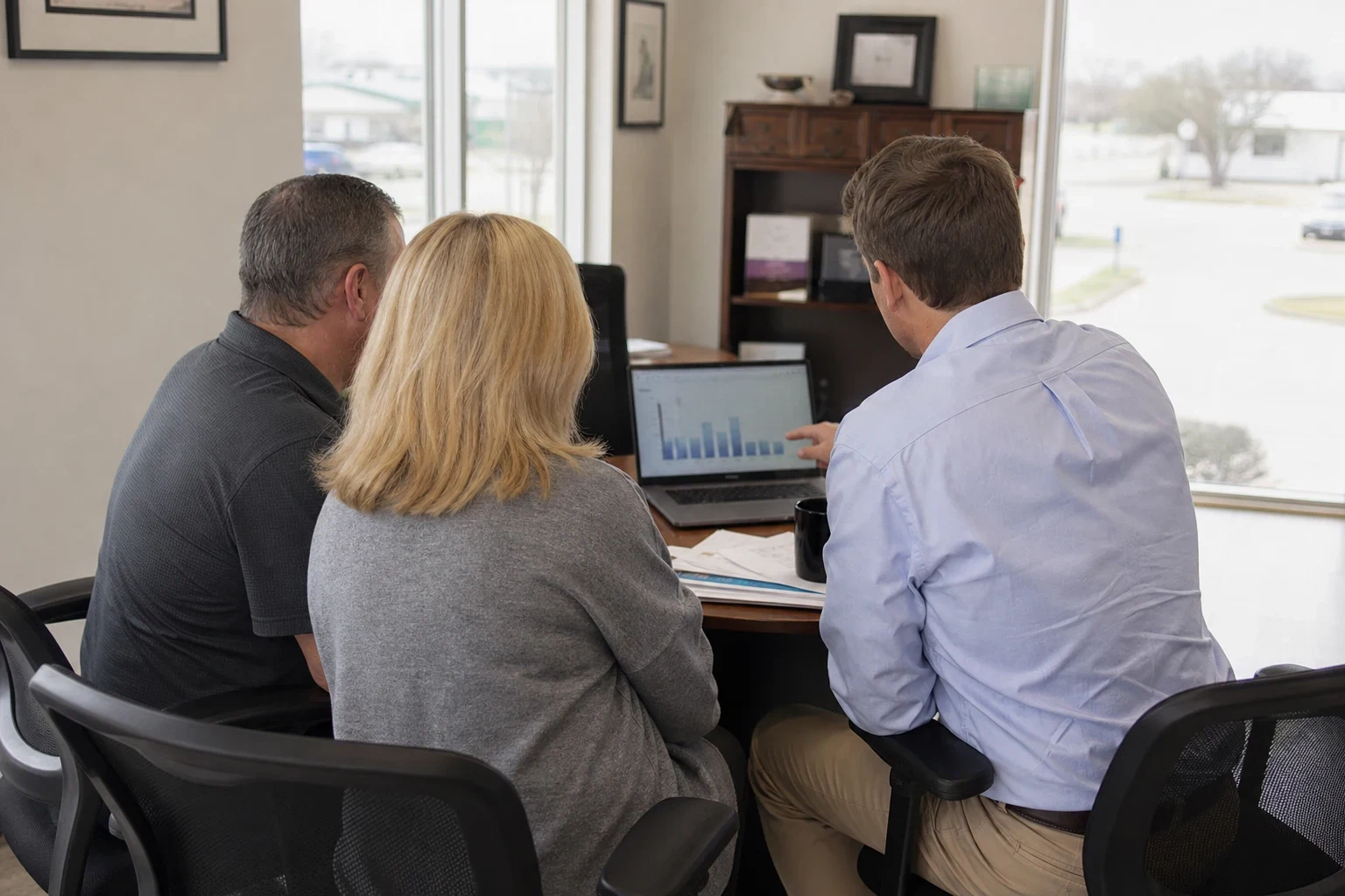 Agent in a light blue button-down points at a bar chart on a laptop while a man and a blonde woman look on at a round office table by large windows on an overcast day.