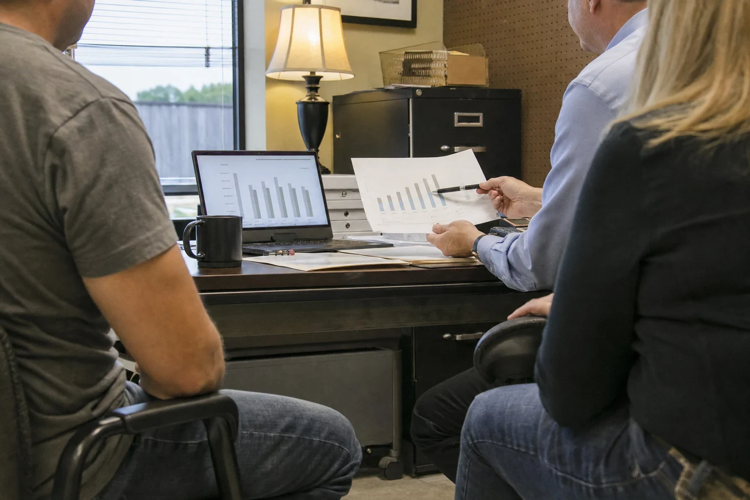Man in a blue shirt points a pen at a printed bar chart while two people sit beside him at a desk with a laptop, lamp and filing cabinet in a small office